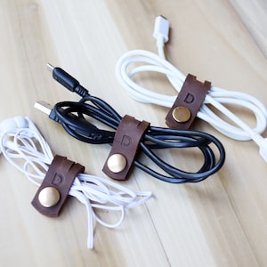 May include: Three brown leather cord organisers with a gold button and the letter "D" embossed on each. The organisers are holding a white cord, a black cord, and a white headphone cord.