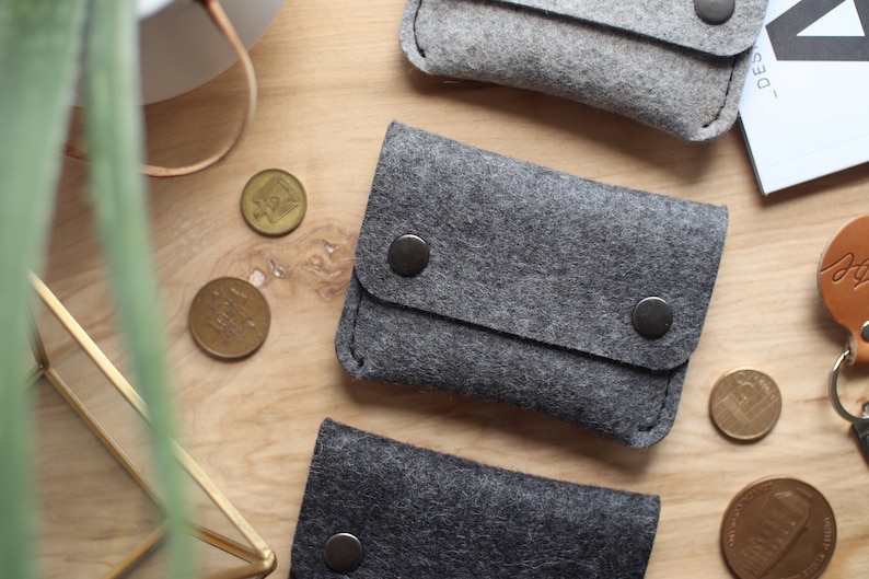 May include: Three gray felt coin purses with a flap closure and a snap button. The purses are on a wooden surface with some coins scattered around.