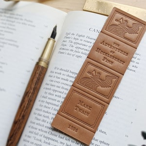 May include: A brown leather bookmark with the title "The Adventures of Huckleberry Finn" by Mark Twain, published in 1884. The bookmark features a decorative design of a bird flying over a wave.