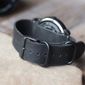 K&ouml;nnte beinhalten: Nahaufnahme eines schwarzen Leder-Uhrenarmbands mit schwarzer Schnalle. Das Armband ist dick und hat eine strukturierte Oberfl&auml;che. Das Zifferblatt der Uhr ist im Hintergrund sichtbar.