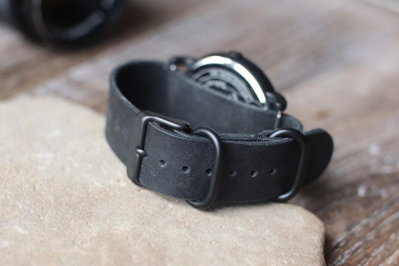 K&ouml;nnte beinhalten: Nahaufnahme eines schwarzen Leder-Uhrenarmbands mit schwarzer Schnalle. Das Armband ist dick und hat eine strukturierte Oberfl&auml;che. Das Zifferblatt der Uhr ist im Hintergrund sichtbar.