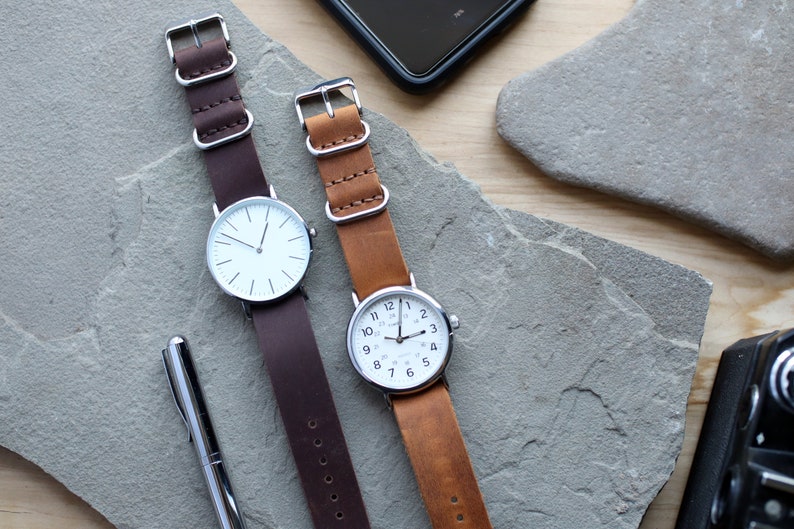 K&ouml;nnte beinhalten: Zwei Armbanduhren mit Lederarmb&auml;ndern. Eine Uhr hat ein dunkelbraunes Armband und ein wei&szlig;es Zifferblatt mit minimalistischen Markierungen. Die andere Uhr hat ein hellbraunes Armband und ein wei&szlig;es Zifferblatt mit schwarzen Ziffern. Beide Uhren haben silberfarbene Metallgeh&auml;use.