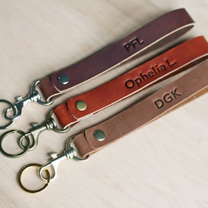May include: Three leather keychains with metal key rings. The keychains are brown, red, and tan. The keychains are personalised with the initials "PFL", "Ophelia L", and "DGK".