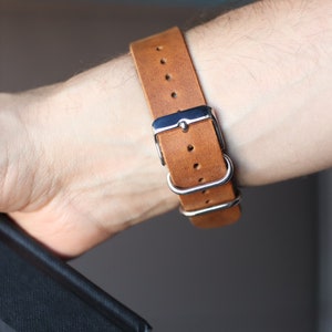 K&ouml;nnte beinhalten: Nahaufnahme eines braunen Lederarmbands mit silberner Schnalle und Schlaufen. Das Armband ist an einem Handgelenk befestigt. Das Armband besteht aus hellbraunem Leder.