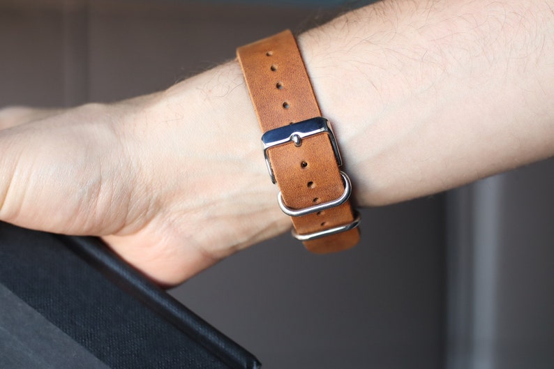 K&ouml;nnte beinhalten: Nahaufnahme eines braunen Lederarmbands mit silberner Schnalle und Schlaufen. Das Armband ist an einem Handgelenk befestigt. Das Armband besteht aus hellbraunem Leder.