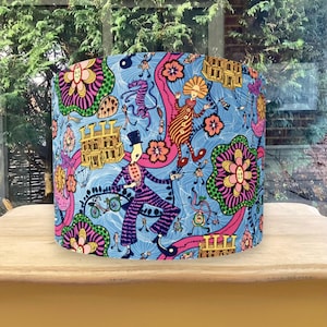 K&ouml;nnte beinhalten: Zylindrischer Lampenschirm mit einem lebendigen, skurrilen Design. Der Lampenschirm hat einen hellblauen Hintergrund mit farbenfrohen Illustrationen von Figuren, Blumen und architektonischen Elementen in Pink, Orange und Gr&uuml;n. Der Lampenschirm steht auf einem Holztisch.