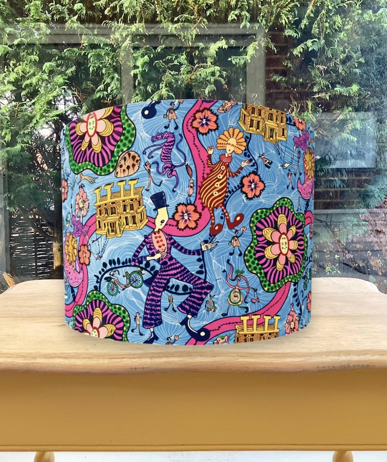 K&ouml;nnte beinhalten: Zylindrischer Lampenschirm mit einem lebendigen, skurrilen Design. Der Lampenschirm hat einen hellblauen Hintergrund mit farbenfrohen Illustrationen von Figuren, Blumen und architektonischen Elementen in Pink, Orange und Gr&uuml;n. Der Lampenschirm steht auf einem Holztisch.