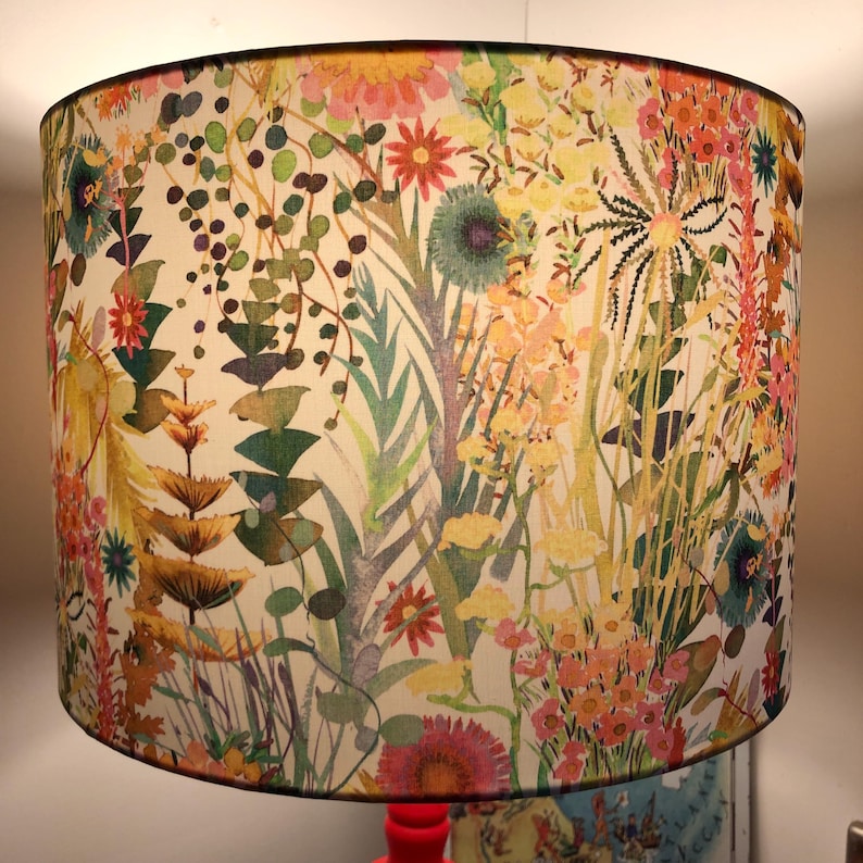 K&ouml;nnte beinhalten: Zylindrischer Lampenschirm mit einem lebendigen Blumenmuster. Das Design umfasst eine Vielzahl farbenfroher Blumen und Bl&auml;tter in Rosa-, Gelb-, Orange- und Gr&uuml;nt&ouml;nen auf cremefarbenem Hintergrund. Der Lampenschirm hat eine schwarze Umrandung.