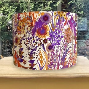 K&ouml;nnte beinhalten: Zylindrischer Lampenschirm mit einem lebendigen Blumenmuster. Das Design zeigt lila, orange und gelbe Blumen und Bl&auml;tter auf cremefarbenem Hintergrund. Ein dekoratives Beleuchtungselement.