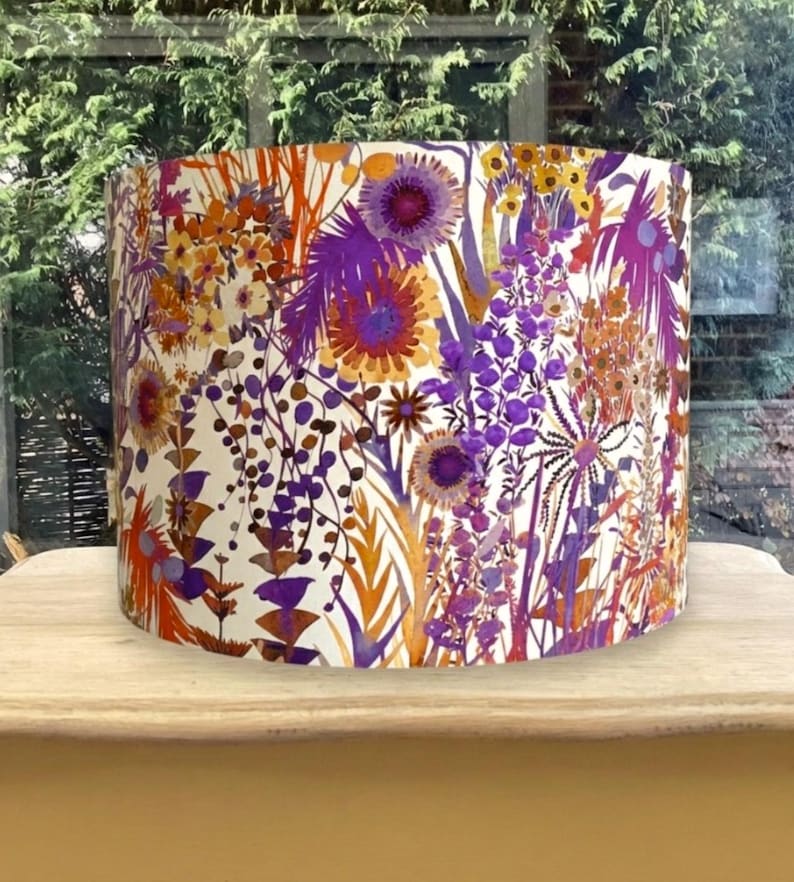 K&ouml;nnte beinhalten: Zylindrischer Lampenschirm mit einem lebendigen Blumenmuster. Das Design zeigt lila, orange und gelbe Blumen und Bl&auml;tter auf cremefarbenem Hintergrund. Ein dekoratives Beleuchtungselement.