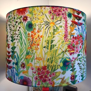 K&ouml;nnte beinhalten: Zylindrischer Lampenschirm mit einem lebendigen Blumenmuster. Das Design zeigt eine Vielzahl farbenfroher Blumen und Bl&auml;tter in Rot-, Gelb-, Orange- und Gr&uuml;nt&ouml;nen auf wei&szlig;em Hintergrund. Ein dekoratives Beleuchtungselement.
