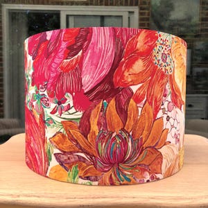K&ouml;nnte beinhalten: Ein zylindrischer Lampenschirm mit einem auff&auml;lligen Blumenmuster. Das Design zeigt gro&szlig;e, stilisierte Blumen in Rot-, Orange- und Rosat&ouml;nen vor einem wei&szlig;en Hintergrund. Der Lampenschirm steht auf einer hellen Holzoberfl&auml;che.