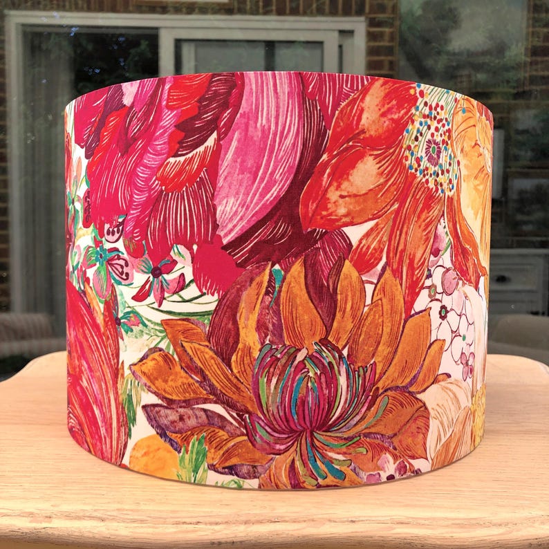 K&ouml;nnte beinhalten: Ein zylindrischer Lampenschirm mit einem auff&auml;lligen Blumenmuster. Das Design zeigt gro&szlig;e, stilisierte Blumen in Rot-, Orange- und Rosat&ouml;nen vor einem wei&szlig;en Hintergrund. Der Lampenschirm steht auf einer hellen Holzoberfl&auml;che.