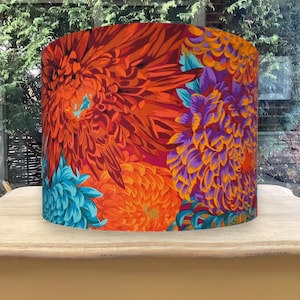 K&ouml;nnte beinhalten: Zylindrischer Lampenschirm mit einem lebendigen Blumenmuster. Das Design zeigt gro&szlig;e, stilisierte Blumen in Orange-, Rot-, Lila- und T&uuml;rkist&ouml;nen. Der Lampenschirm steht auf einem hellen Holztisch, mit einem verschwommenen Hintergrund im Freien.