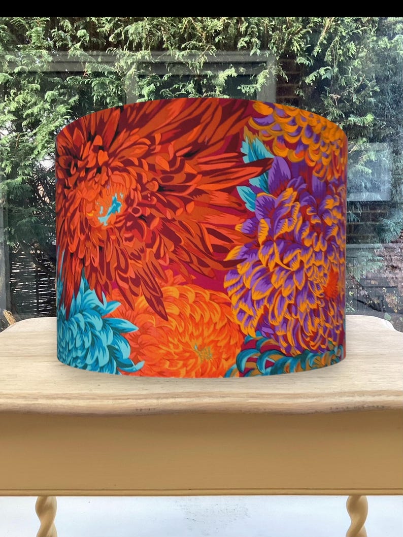 K&ouml;nnte beinhalten: Zylindrischer Lampenschirm mit einem lebendigen Blumenmuster. Das Design zeigt gro&szlig;e, stilisierte Blumen in Orange-, Rot-, Lila- und T&uuml;rkist&ouml;nen. Der Lampenschirm steht auf einem hellen Holztisch, mit einem verschwommenen Hintergrund im Freien.