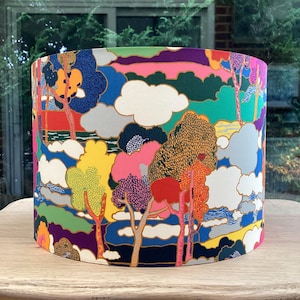 K&ouml;nnte beinhalten: Ein zylindrischer Lampenschirm mit einem farbenfrohen Landschaftsdesign. Das Muster zeigt B&auml;ume, Wolken und eine Vielzahl von Farben, darunter Blau, Gr&uuml;n, Rosa, Gelb und Orange. Der Lampenschirm steht auf einer Holzoberfl&auml;che.
