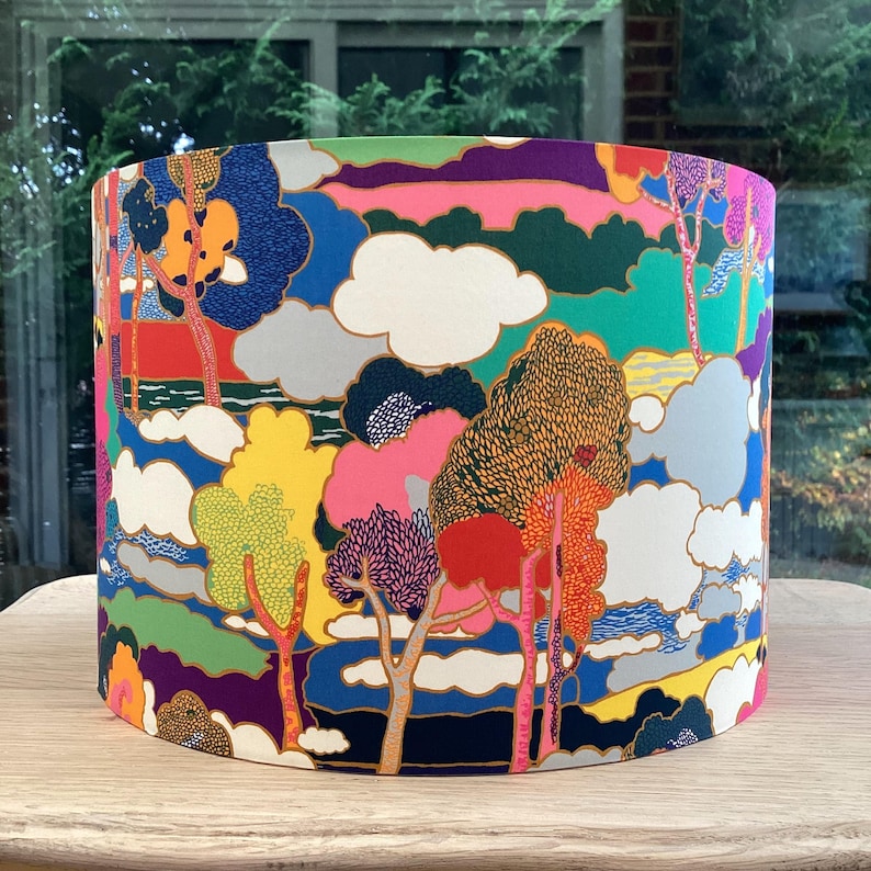 K&ouml;nnte beinhalten: Ein zylindrischer Lampenschirm mit einem farbenfrohen Landschaftsdesign. Das Muster zeigt B&auml;ume, Wolken und eine Vielzahl von Farben, darunter Blau, Gr&uuml;n, Rosa, Gelb und Orange. Der Lampenschirm steht auf einer Holzoberfl&auml;che.