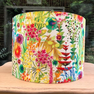 K&ouml;nnte beinhalten: Zylindrischer Lampenschirm mit einem lebendigen Blumenmuster. Das Design zeigt eine Mischung aus bunten Blumen und Bl&auml;ttern in Rot-, Gelb-, Rosa- und Gr&uuml;nt&ouml;nen vor einem wei&szlig;en Hintergrund. Der Lampenschirm steht auf einer Holzoberfl&auml;che.