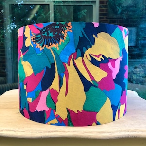 K&ouml;nnte beinhalten: Zylindrischer Lampenschirm mit einem lebendigen Blumenmuster. Das Design zeigt gro&szlig;e, stilisierte Blumen in Gelb-, Rosa-, Blau-, Gr&uuml;n- und Oranget&ouml;nen vor einem dunkelblauen Hintergrund. Der Lampenschirm steht auf einer hellen Oberfl&auml;che.