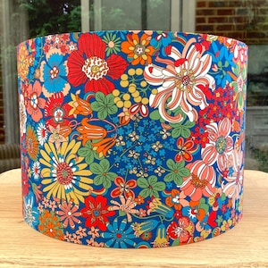 K&ouml;nnte beinhalten: Zylindrischer Lampenschirm mit einem lebendigen Blumenmuster. Das Design zeigt einen tiefblauen Hintergrund, der mit einer Vielzahl farbenfroher Blumen in Rot, Orange, Gelb und Wei&szlig; verziert ist. Der Lampenschirm hat einen Durchmesser von ca. 25 cm und ist 20 cm hoch.