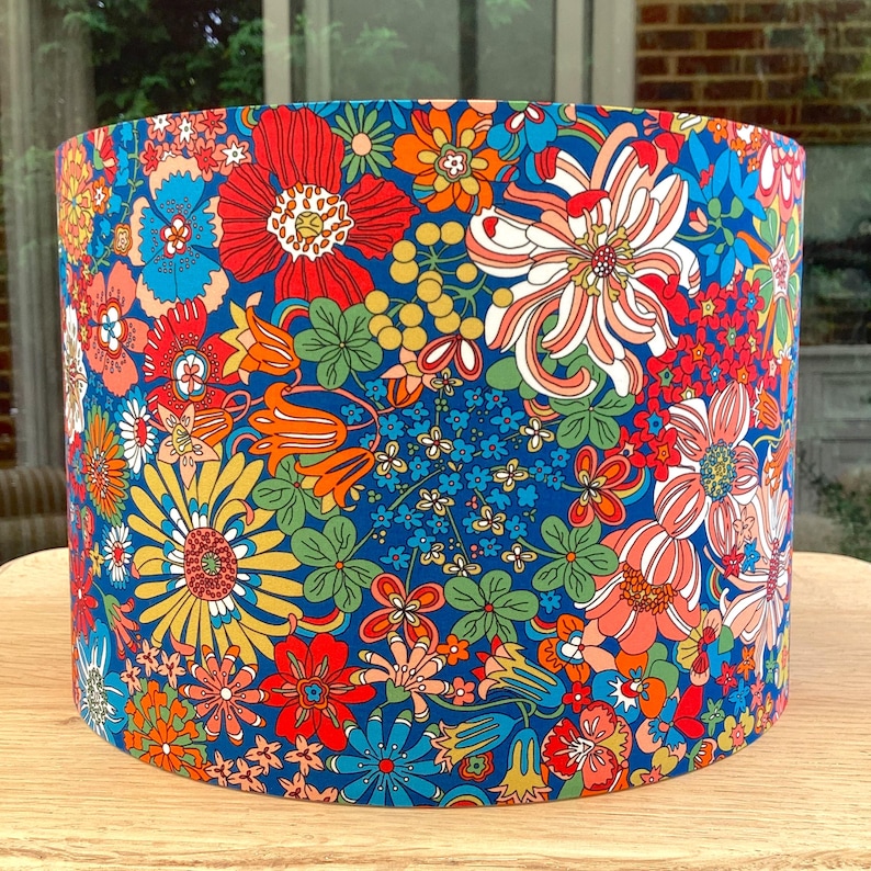 K&ouml;nnte beinhalten: Zylindrischer Lampenschirm mit einem lebendigen Blumenmuster. Das Design zeigt einen tiefblauen Hintergrund, der mit einer Vielzahl farbenfroher Blumen in Rot, Orange, Gelb und Wei&szlig; verziert ist. Der Lampenschirm hat einen Durchmesser von ca. 25 cm und ist 20 cm hoch.