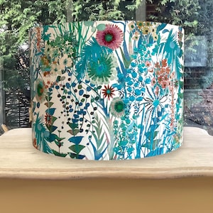 K&ouml;nnte beinhalten: Ein zylindrischer Lampenschirm mit floralem Muster. Das Design zeigt eine Vielzahl von Blumen und Bl&auml;ttern in T&uuml;rkis-, Blau-, Rosa- und Oranget&ouml;nen auf wei&szlig;em Hintergrund. Der Lampenschirm steht auf einem hellen Holztisch.