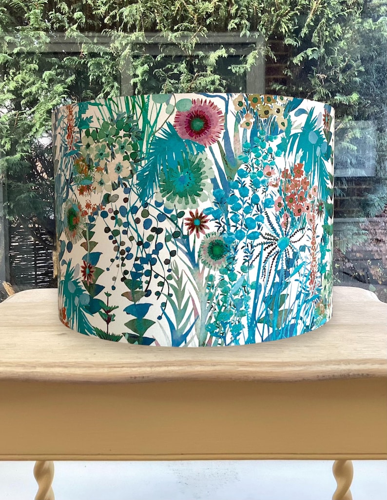 K&ouml;nnte beinhalten: Ein zylindrischer Lampenschirm mit floralem Muster. Das Design zeigt eine Vielzahl von Blumen und Bl&auml;ttern in T&uuml;rkis-, Blau-, Rosa- und Oranget&ouml;nen auf wei&szlig;em Hintergrund. Der Lampenschirm steht auf einem hellen Holztisch.