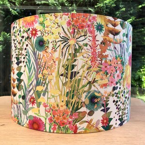 K&ouml;nnte beinhalten: Ein zylindrischer Lampenschirm mit einem lebendigen Blumendesign. Der Lampenschirm zeigt eine Vielzahl farbenfroher Blumen und Bl&auml;tter in Rosa-, Gelb-, Orange- und Gr&uuml;nt&ouml;nen vor einem wei&szlig;en Hintergrund. Das Design erinnert an ein Gartenthema.