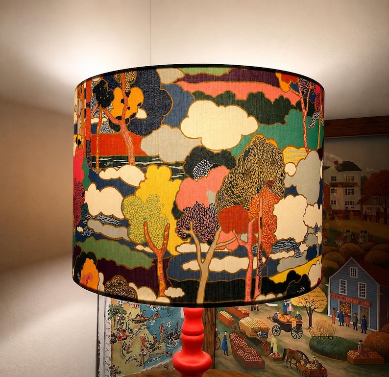 K&ouml;nnte beinhalten: Zylindrischer Lampenschirm mit farbenfrohem Landschaftsdesign. Das Kunstwerk zeigt B&auml;ume, Wolken und eine Vielzahl von Farben, darunter Rot, Gelb, Blau und Gr&uuml;n. Der Lampenschirm ist auf einem roten Lampenfu&szlig; montiert.