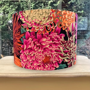 K&ouml;nnte beinhalten: Zylindrischer Lampenschirm mit einem lebendigen Blumenmuster. Das Design zeigt gro&szlig;e, stilisierte Blumen in Rosa-, Orange- und Cremet&ouml;nen vor einem schwarzen Hintergrund. Der Lampenschirm wird auf einem hellen Holztisch pr&auml;sentiert. Der Durchmesser betr&auml;gt ca. 30 cm.