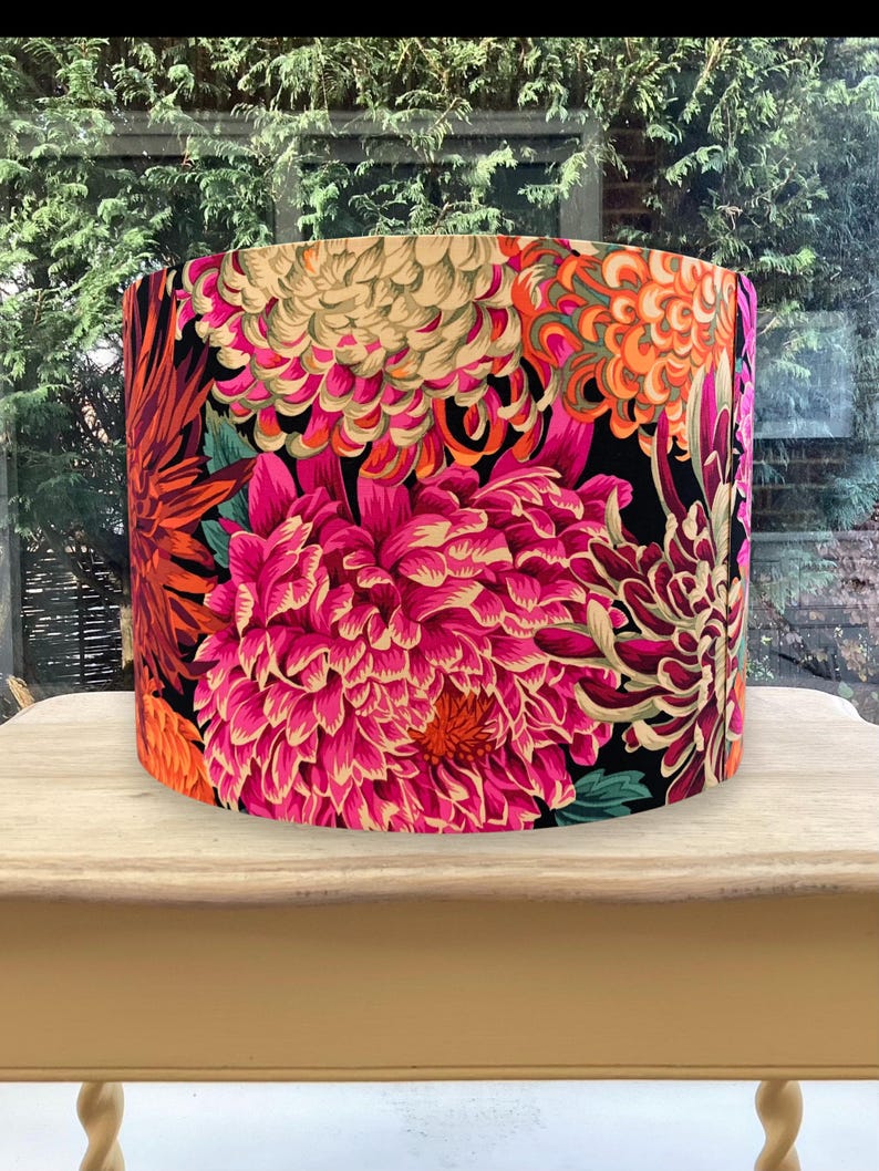 K&ouml;nnte beinhalten: Zylindrischer Lampenschirm mit einem lebendigen Blumenmuster. Das Design zeigt gro&szlig;e, stilisierte Blumen in Rosa-, Orange- und Cremet&ouml;nen vor einem schwarzen Hintergrund. Der Lampenschirm wird auf einem hellen Holztisch pr&auml;sentiert. Der Durchmesser betr&auml;gt ca. 30 cm.