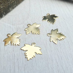 May include: Five gold-coloured maple leaf charms. Each charm has a small loop at the top for attaching to jewellery or other crafts. The leaves have detailed edges and a shiny finish.