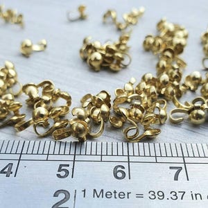 Pu&ograve; includere: Primo piano di un righello che misura un mucchio di chiusure metalliche dorate. Il righello mostra le misure in pollici. Le chiusure sono piccole e hanno una sfera arrotondata a un'estremit&agrave; e un gancio all'altra.