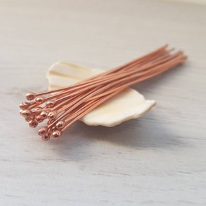 May include: A bunch of copper head pins with rounded ends, arranged on a white seashell. The pins are about 1 inch long.