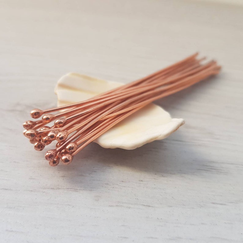 May include: A bunch of copper head pins with rounded ends, arranged on a white seashell. The pins are about 1 inch long.