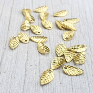 May include: A scattering of small, gold-coloured leaf charms on a light-coloured wooden surface. Each charm features a detailed leaf vein pattern and a small hole for threading. These charms are suitable for jewellery making and other craft projects.