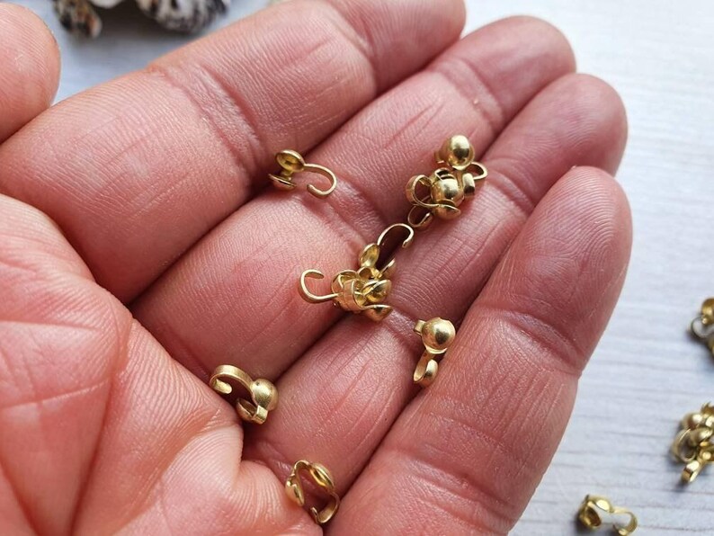 Pu&ograve; includere: Primo piano di una mano che tiene un gruppo di chiusure metalliche dorate. Le chiusure sono piccole e hanno una forma rotonda con un anello su un lato. Sono utilizzate per la creazione di gioielli o altri progetti di artigianato.