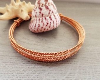 May include: A copper-colored, twisted wire bracelet with three strands. A seashell with red and white markings and a beige starfish are in the background. The bracelet is on a light gray surface.
