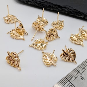 May include: Gold-tone leaf-shaped earring findings with a post and a loop. The findings are scattered on a white surface with a ruler in the background.