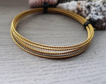 May include: A stack of four gold-colored, braided bracelets. The bracelets are held together with a black band. The background is a light gray wooden surface with a few decorative items.
