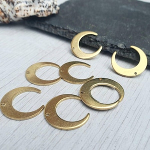 May include: Six gold-toned metal crescent moon charms with a hole in the centre. The charms are arranged in a semi-circle on a light wood surface.