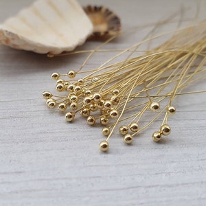 Peut inclure: Un ensemble d'épingles à tête perlée dorées, chacune avec une petite perle ronde au bout d'un fil fin. Les épingles sont regroupées, suggérant leur utilisation dans l'artisanat ou la bijouterie. Un coquillage est en arrière-plan.