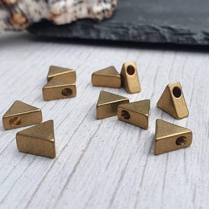May include: A group of ten gold-coloured triangle-shaped metal beads with a hole in the centre of each bead. The beads are arranged on a light wood surface.