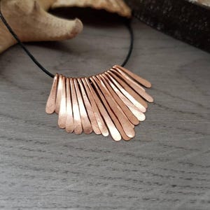 May include: A copper necklace with a fan-shaped pendant made of multiple thin, hammered copper strips. The necklace is on a black cord.