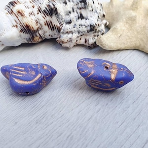 May include: Two purple ceramic beads with gold accents. The beads are shaped like birds with detailed carvings.
