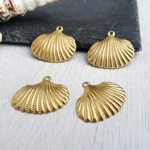 May include: Four gold-coloured seashell charms with a textured, ridged design. Each charm has a small loop at the top for attaching to jewellery or other crafts. The charms are arranged on a light-coloured wooden surface.