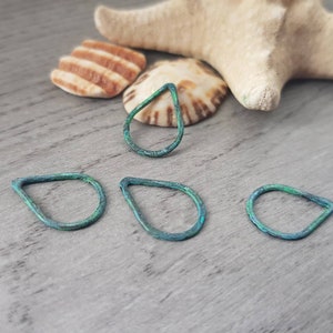May include: Four teal teardrop-shaped metal jewellery components. The components have a textured surface and are arranged on a grey wooden surface, with seashells and a starfish in the background.