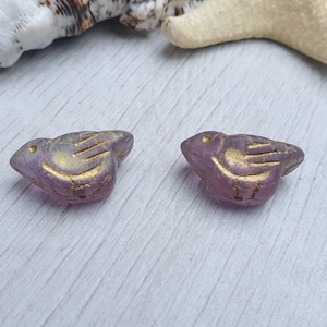 May include: Two purple bird-shaped beads with gold accents. The beads have a textured surface and are set against a light-colored wooden background. The bird beads are a decorative element, possibly for jewelry or crafts.