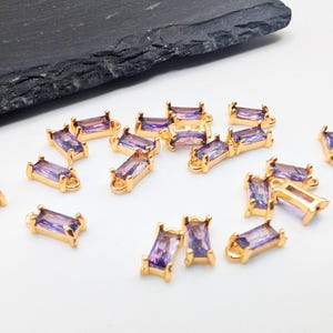 May include: A group of 18 gold-toned rectangular charms with purple iridescent glass stones. The charms have a loop at the top for attaching to jewelry.
