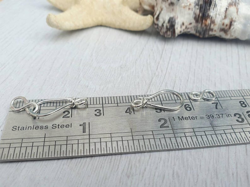 Może przedstawiać: Dwa srebrne metalowe zapięcia, każde o długości około 2,5 cm, pokazane są na linijce z oznaczeniami w calach i centymetrach. Linijka zawiera napis "Stainless Steel" i "1 Meter = 39.37 in".